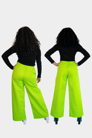 Back view of Aisha (left) and Haydon (right) wearing the Dolores Pantacourt in lime green. Cotton twill wide-legged pants with elastic waistband and invisible zipper, with pockets and belt rings. 100% Brazilian grown BCI cotton. Ethically produced in downtown São Paulo, Brazil